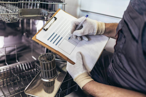 Male Technician Sitting Near Dishwasher Writing On Clipboard In Kitchen, close up.